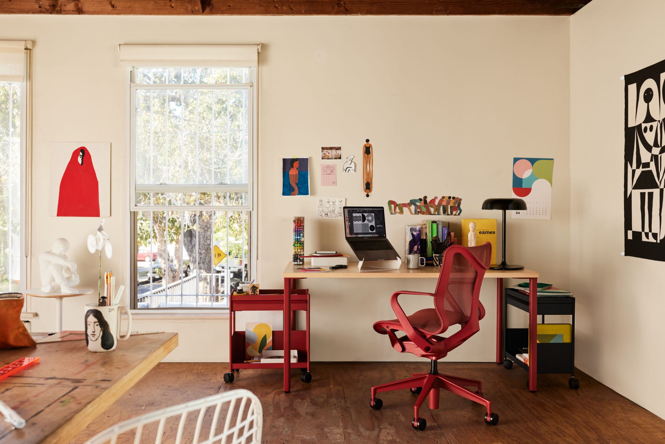 Espaço de trabalho criativo com cadeira ergonômica vermelha, mesa de escritório, notebook e elementos de decoração artística em ambiente iluminado naturalmente.