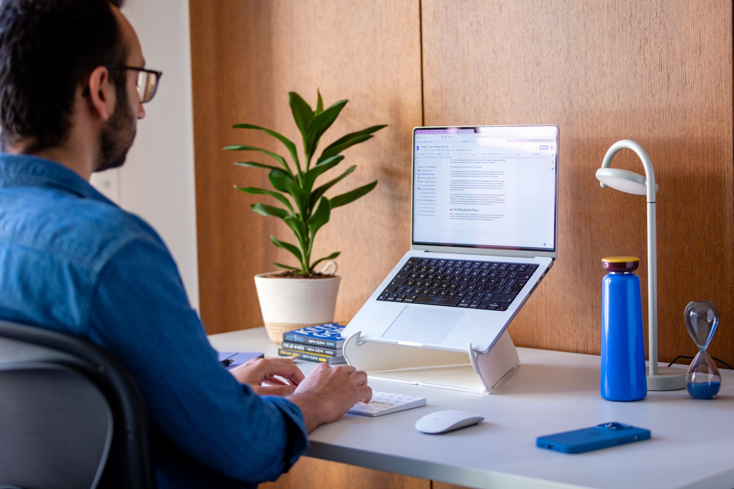 Profissional trabalhando em estação de home office com notebook elevado em suporte ergonômico, teclado externo e ambiente organizado.