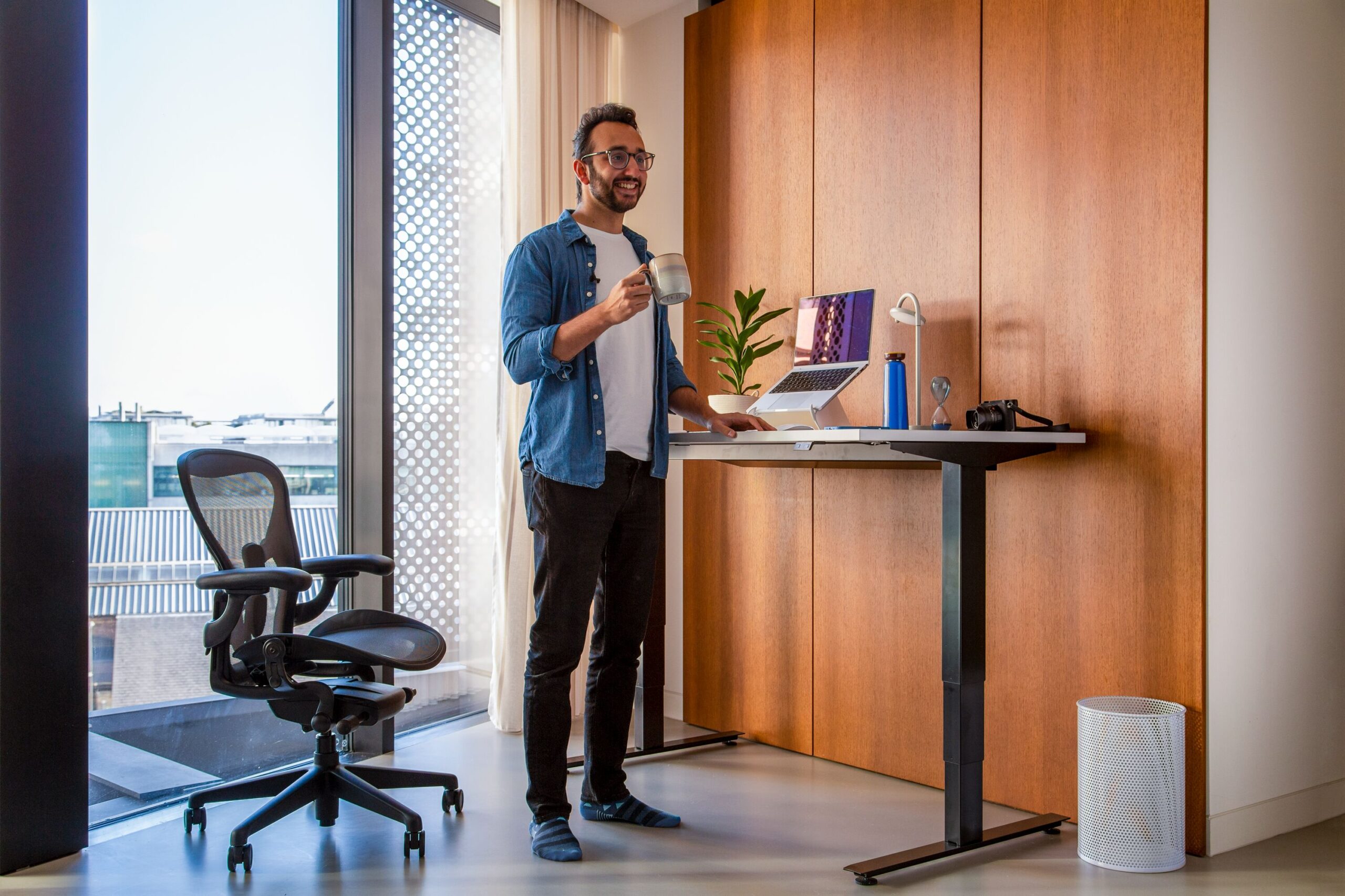 Homem sorri enquanto segura uma xícara de café ao lado de sua standing desk