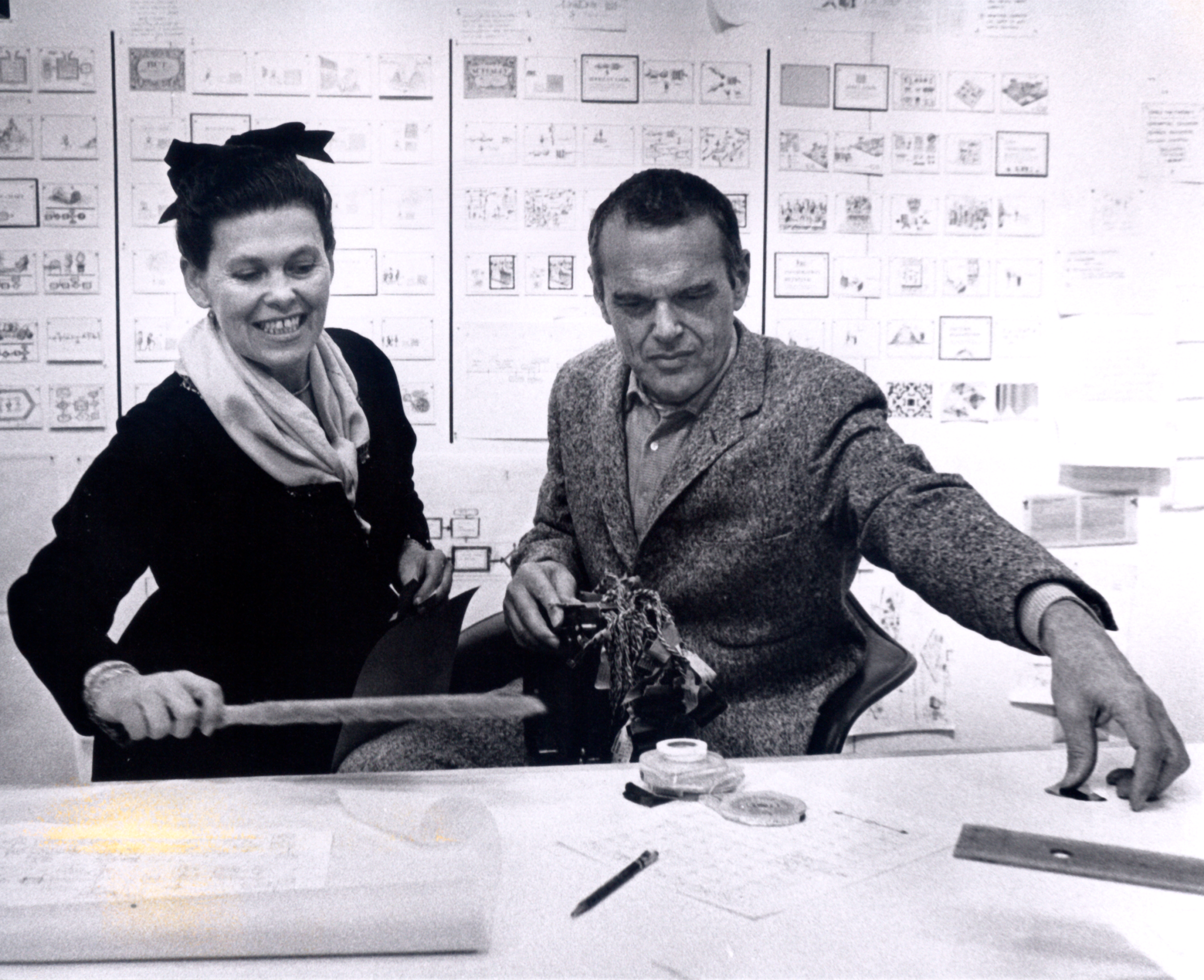 Fotografia histórica de dois designers trabalhando em projetos e protótipos em estúdio de design, cercados por painéis de planejamento e esboços.