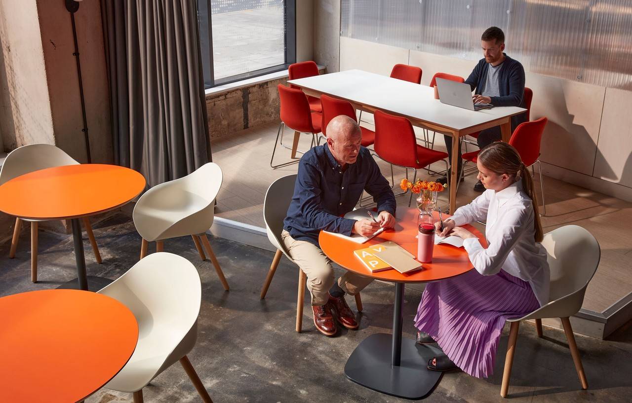 casal de colegas de trabalho sentados em uma mesa laranja com cadeiras eames