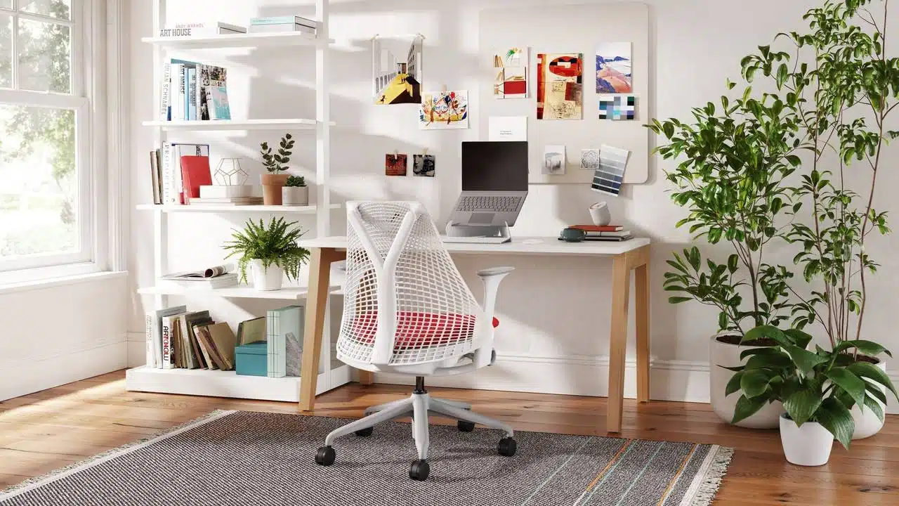 Home office com design inspirador, composto por uma mesa branca com pernas de madeira, cadeira ergonômica branca com almofada vermelha e prateleira decorativa repleta de livros e plantas. O ambiente conta com um painel de inspiração na parede, laptop, luminária e grandes plantas em vasos, iluminado por luz natural que entra pela janela, criando um espaço organizado e acolhedor.